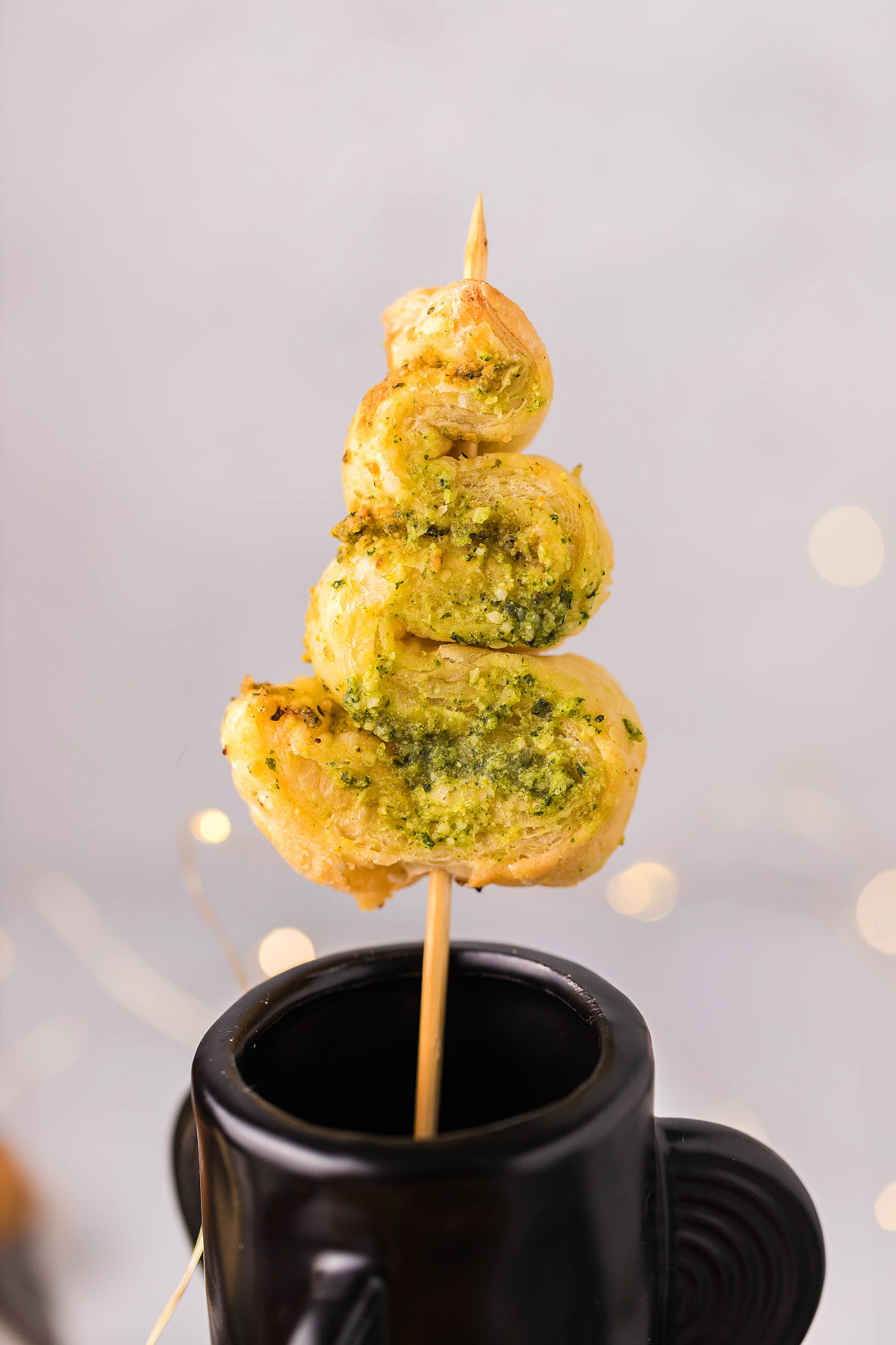 A spiral puff pastry with green pesto on a skewer, placed in a black cup—these Pesto Puff Pastry Christmas Trees make festive and delicious Christmas appetizers.
