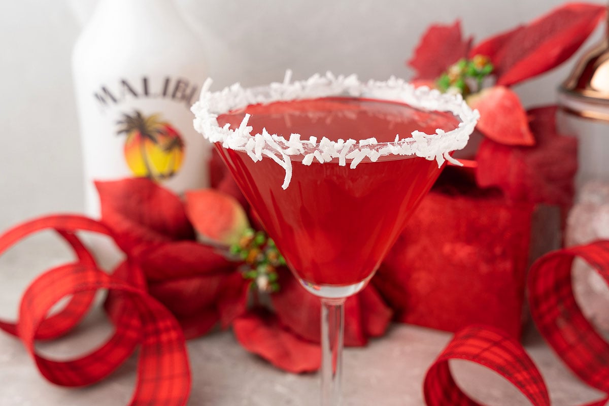 A Red Christmas Cocktail in a martini glass with a coconut rim, surrounded by festive red decorations and a Malibu bottle—this Santa’s Hat Coconut Rum Cocktail is the perfect holiday drink.