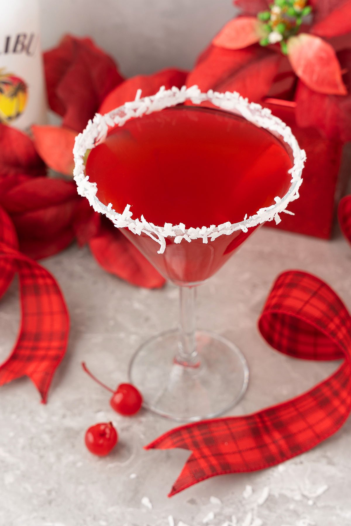 Red christmas cocktail in a martini glass with a coconut rim, garnished with cherries and surrounded by festive holiday decor—this Santa’s Hat Coconut Rum Cocktail is the perfect holiday drink.