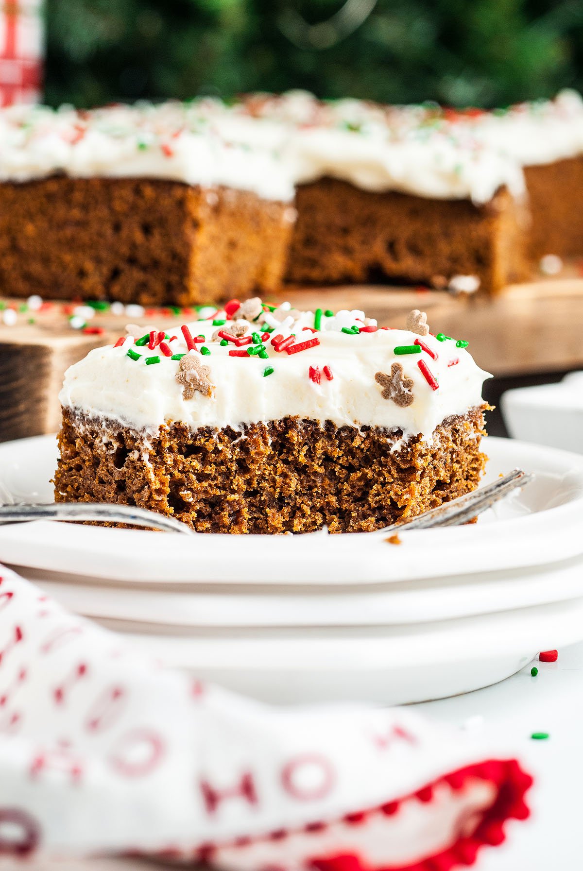 A slice of old fashioned gingerbread cake with white frosting and holiday sprinkles sits on a plate with a fork, perfect as a fall or holiday dessert.