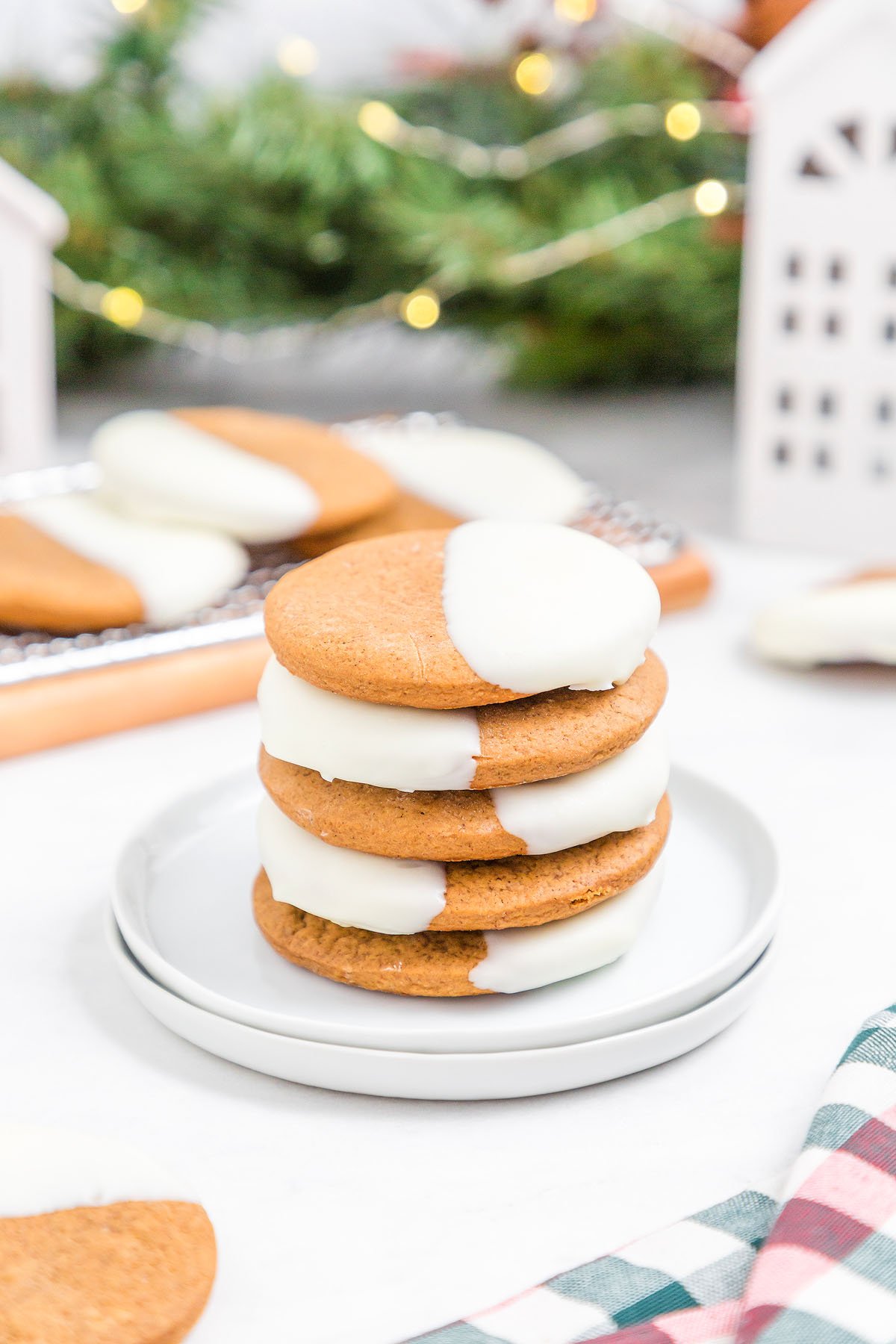 A stack of round white chocolate dipped ginger cookies half-dipped in white icing on a white plate, with holiday decor in the background.