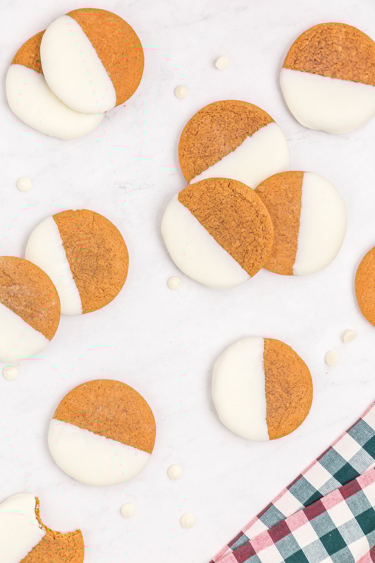 Round gingerbread white chocolate cookies are half-dipped in creamy white chocolate, arranged on a white surface with a checkered cloth in the corner.