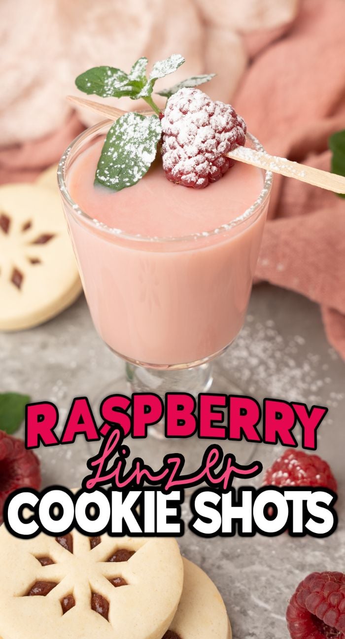 A pink raspberry drink in a shot glass, topped with a raspberry and leaf, surrounded by cookies—perfect as Raspberry Linzer Cookie Shots for your next holiday cocktail or Christmas shots recipe.