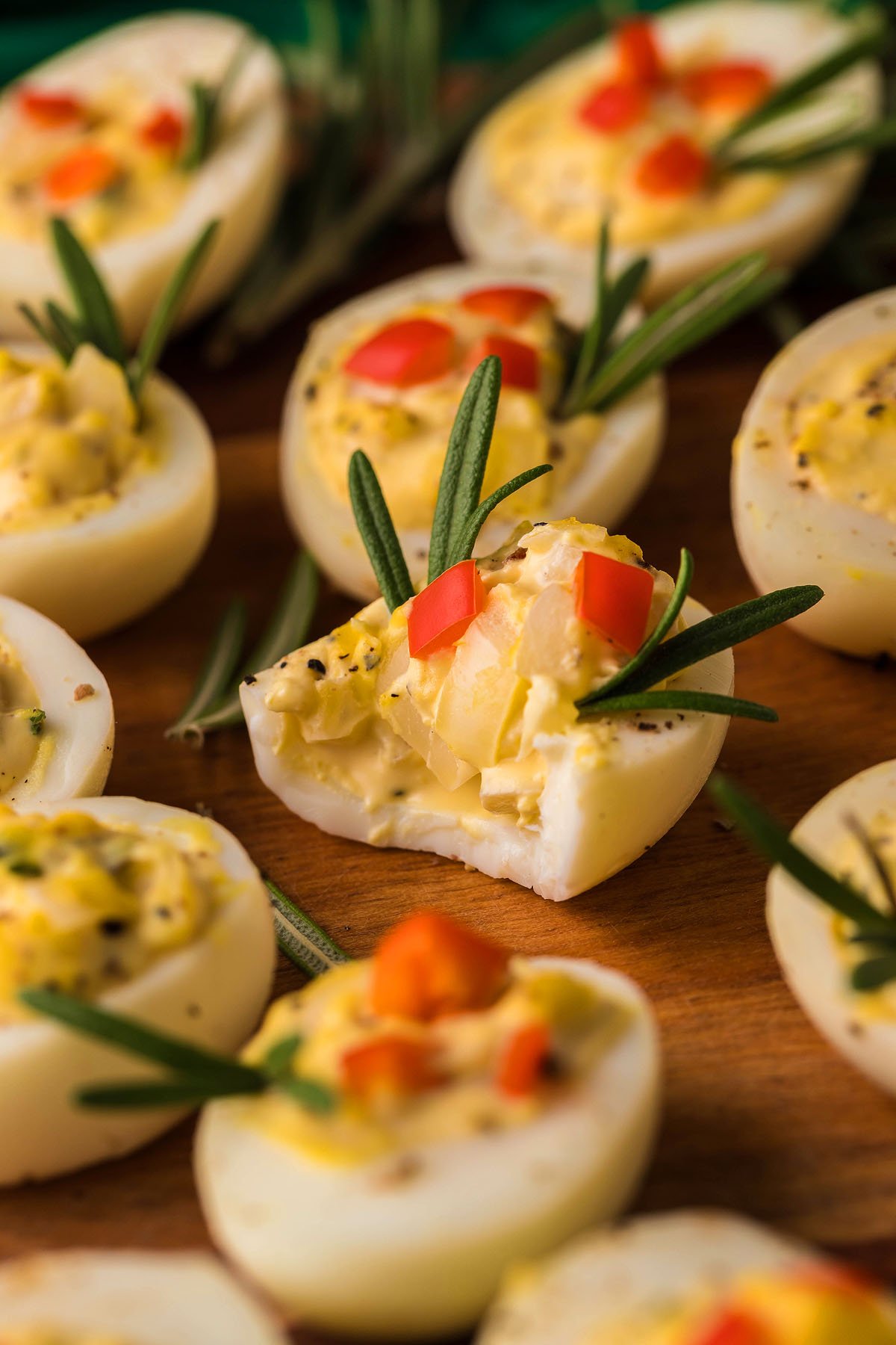 Christmas Deviled Eggs garnished with rosemary sprigs and small diced red peppers make a festive holiday appetizer, perfect for your Christmas party spread. Set on a rustic wooden surface, they bring cheer to any gathering.