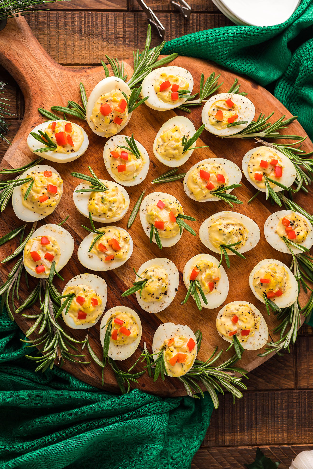 Deviled eggs garnished with herbs and diced red peppers, arranged on a round wooden board with rosemary sprigs—perfect as a festive holiday appetizer for your Christmas party.