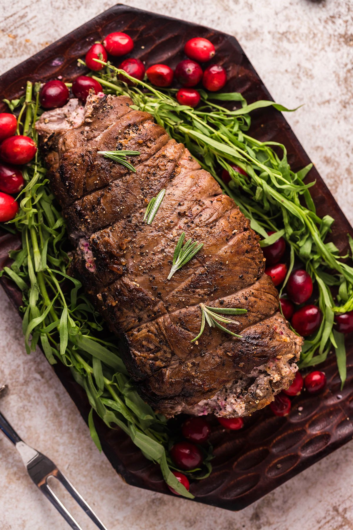 A cooked roast beef garnished with rosemary, surrounded by cranberries and green herbs on a dark platter—perfect inspiration for a Cranberry Cream Cheese Stuffed Flank Steak or a festive Christmas steak recipe.