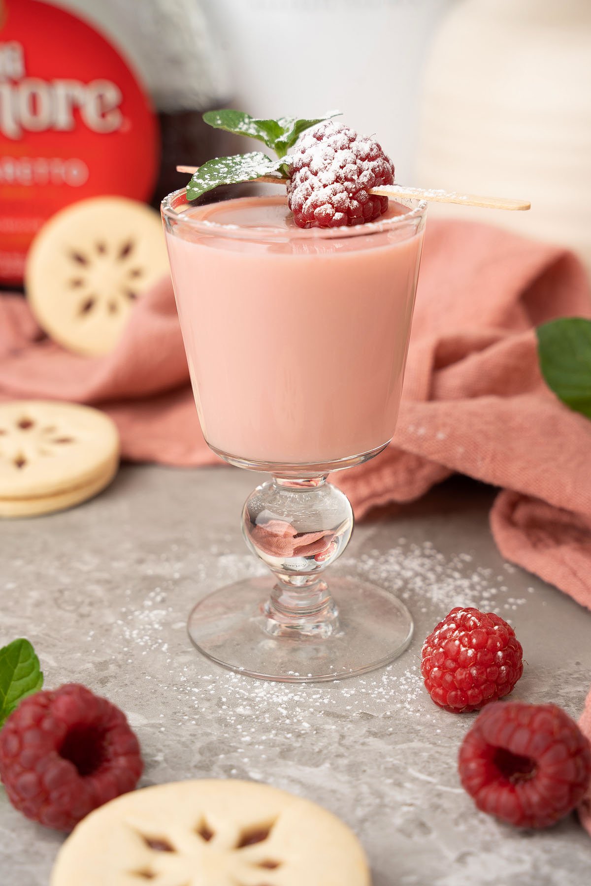 A pink creamy drink in a glass, garnished with raspberries, mint, and powdered sugar, paired with cookies—perfect for serving as festive Raspberry Linzer Cookie Shots at your next holiday cocktail party.