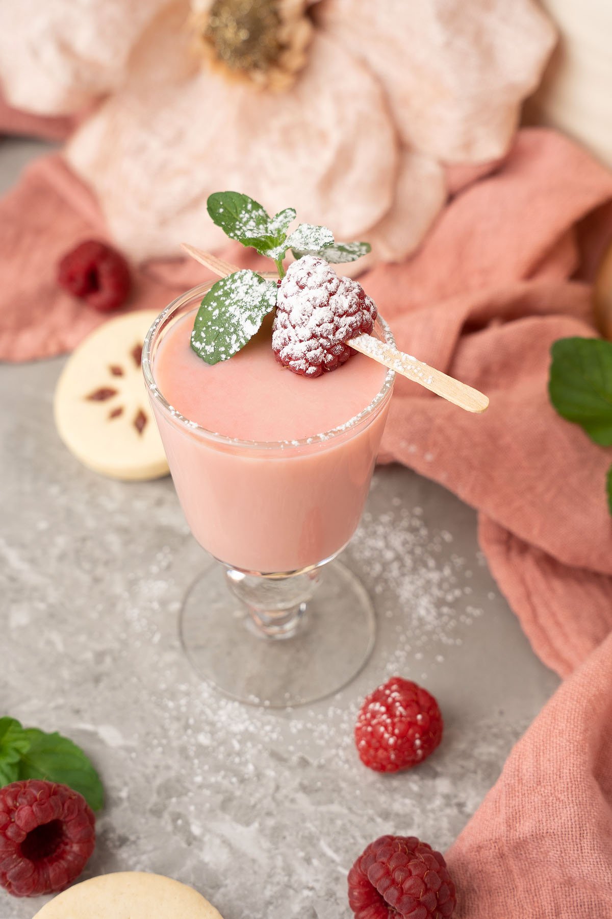 Pink smoothie in a glass, garnished with raspberry, mint, and powdered sugar, surrounded by cookies and raspberries—perfectly capturing the festive charm of a Raspberry Linzer Cookie Shots holiday cocktail.
