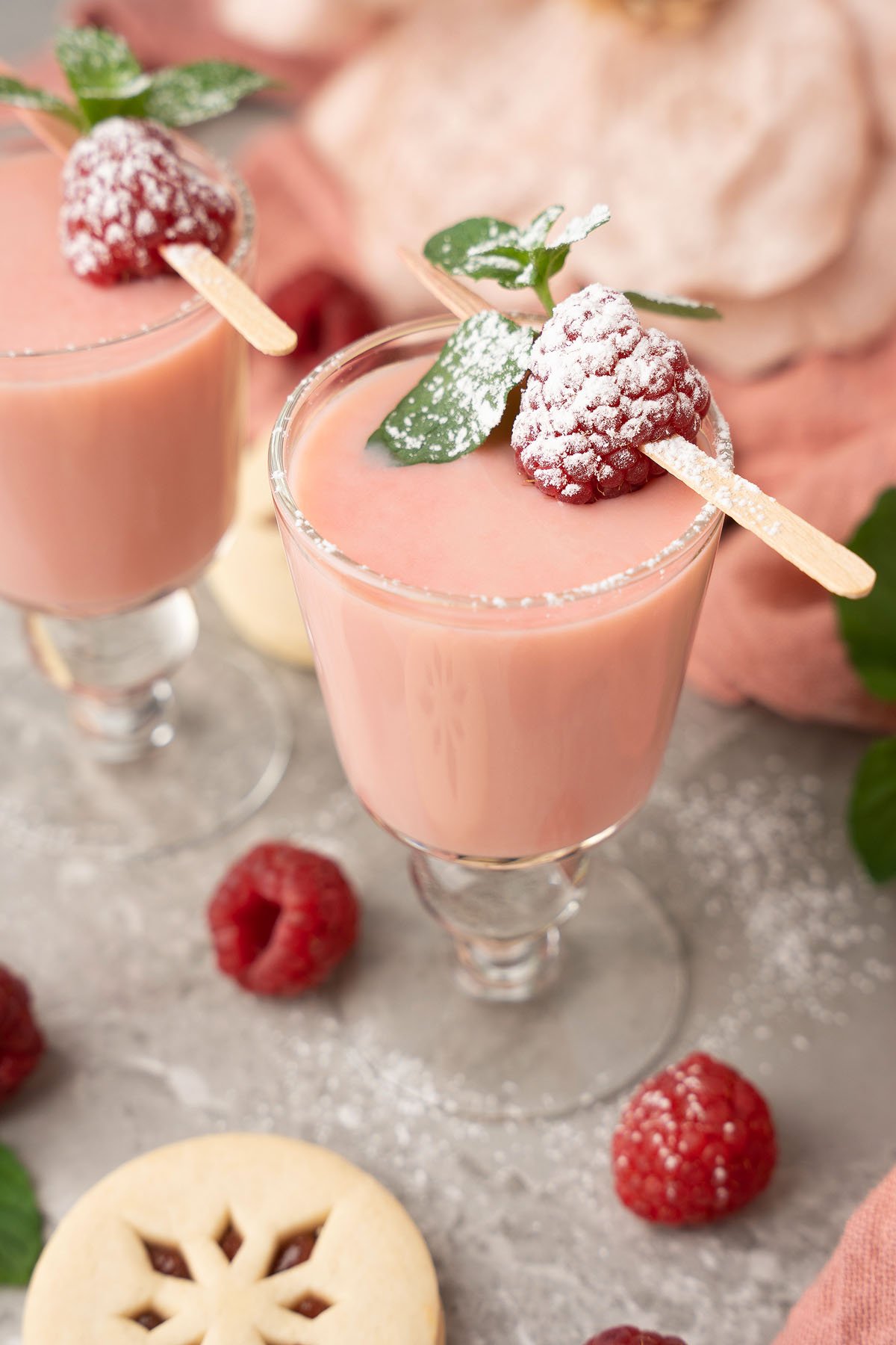 A glass of pink mousse topped with a raspberry, mint leaf, and powdered sugar, surrounded by raspberries—perfect for pairing with Raspberry Linzer Cookie Shots for a festive holiday cocktail experience.