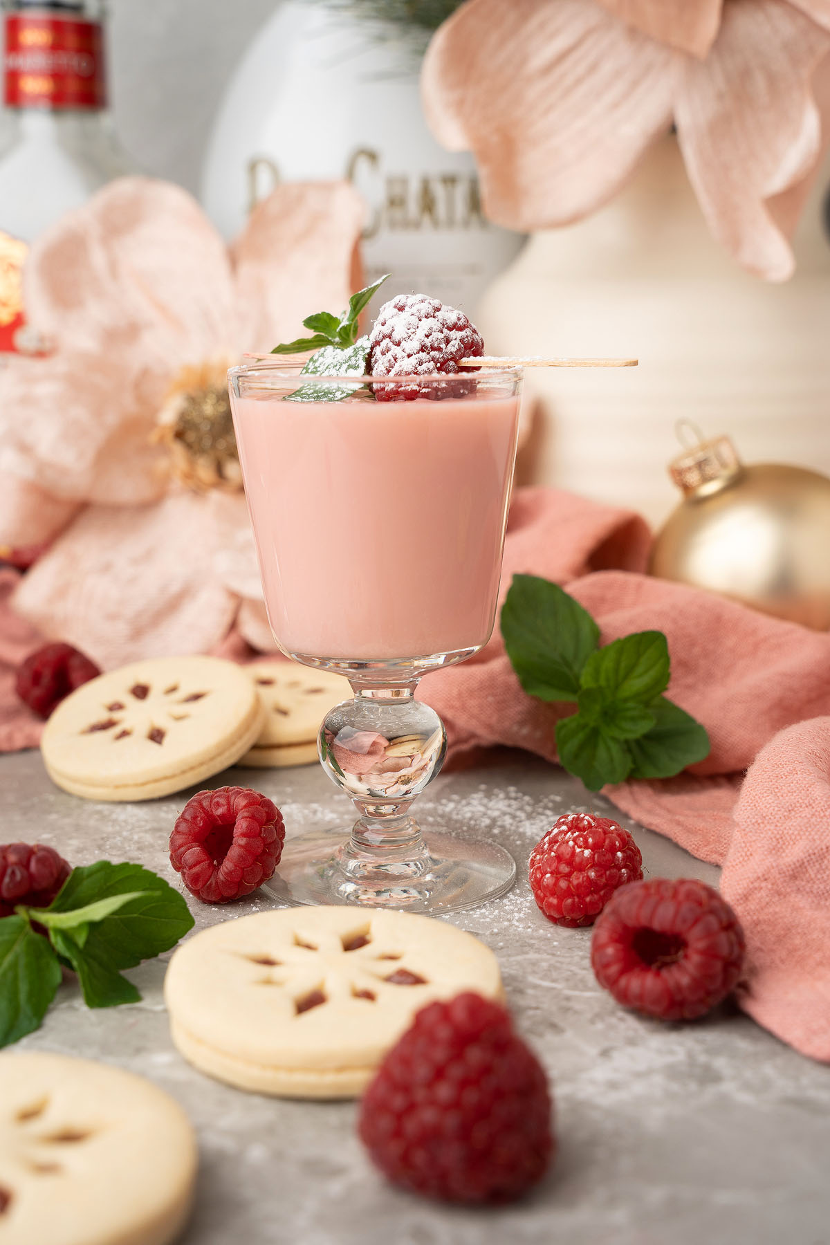 A glass of pink raspberry drink, garnished with raspberries and mint, sits beside cookies and fresh berries—perfect for serving as Raspberry Linzer Cookie Shots or a festive holiday cocktail.