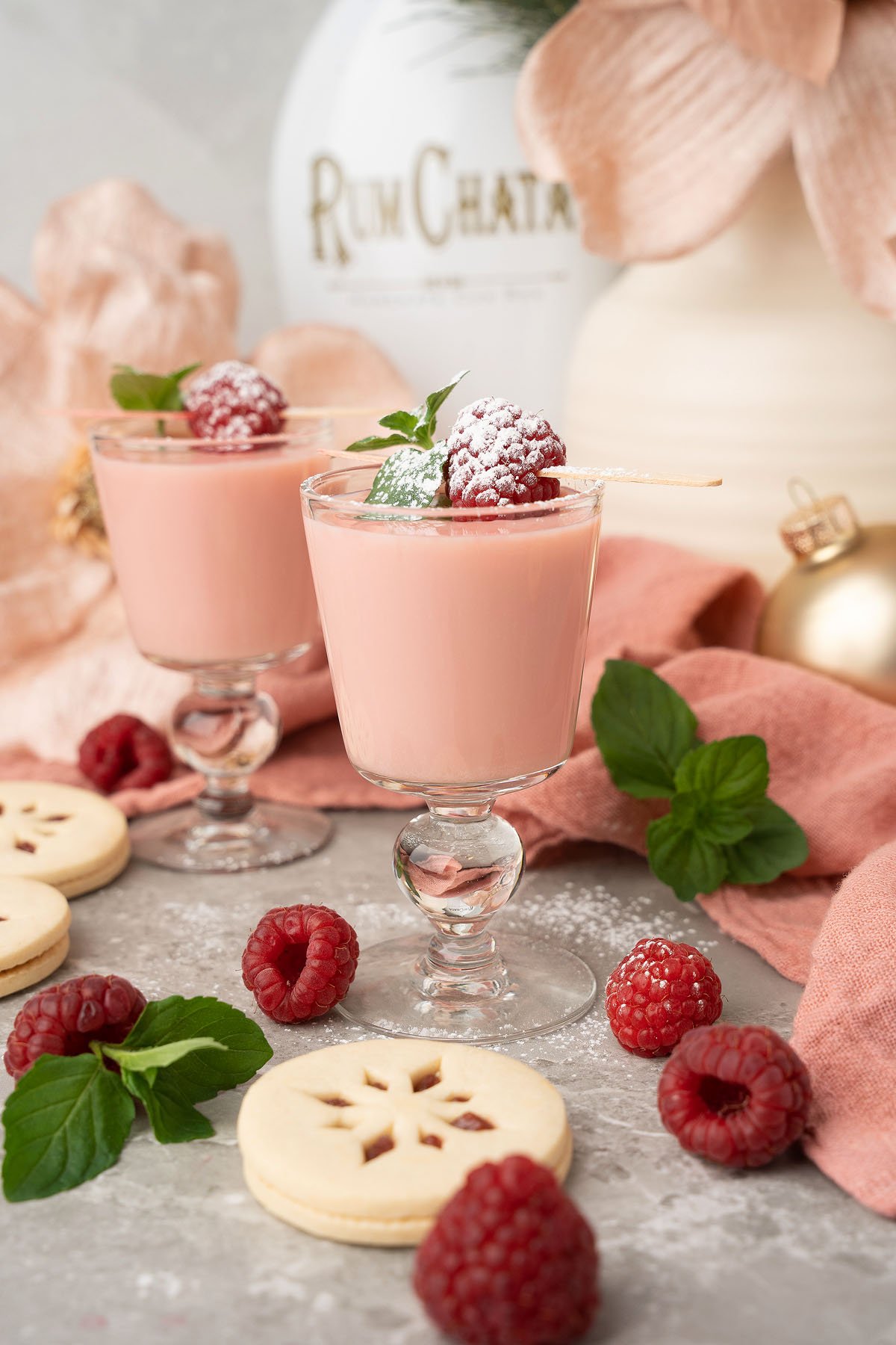 Two glasses of pink creamy Raspberry Linzer Cookie Shots garnished with raspberries and mint, surrounded by cookies and fresh raspberries—perfect for a festive holiday cocktail treat.