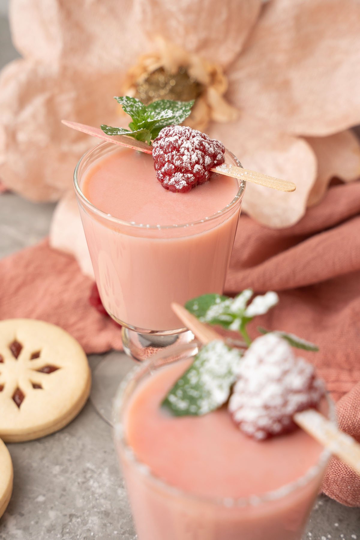 Two glasses of pink mousse topped with raspberries, mint, and powdered sugar sit beside cookies and a flower—perfect alongside festive treats like Raspberry Linzer Cookie Shots or as a sweet holiday cocktail dessert.