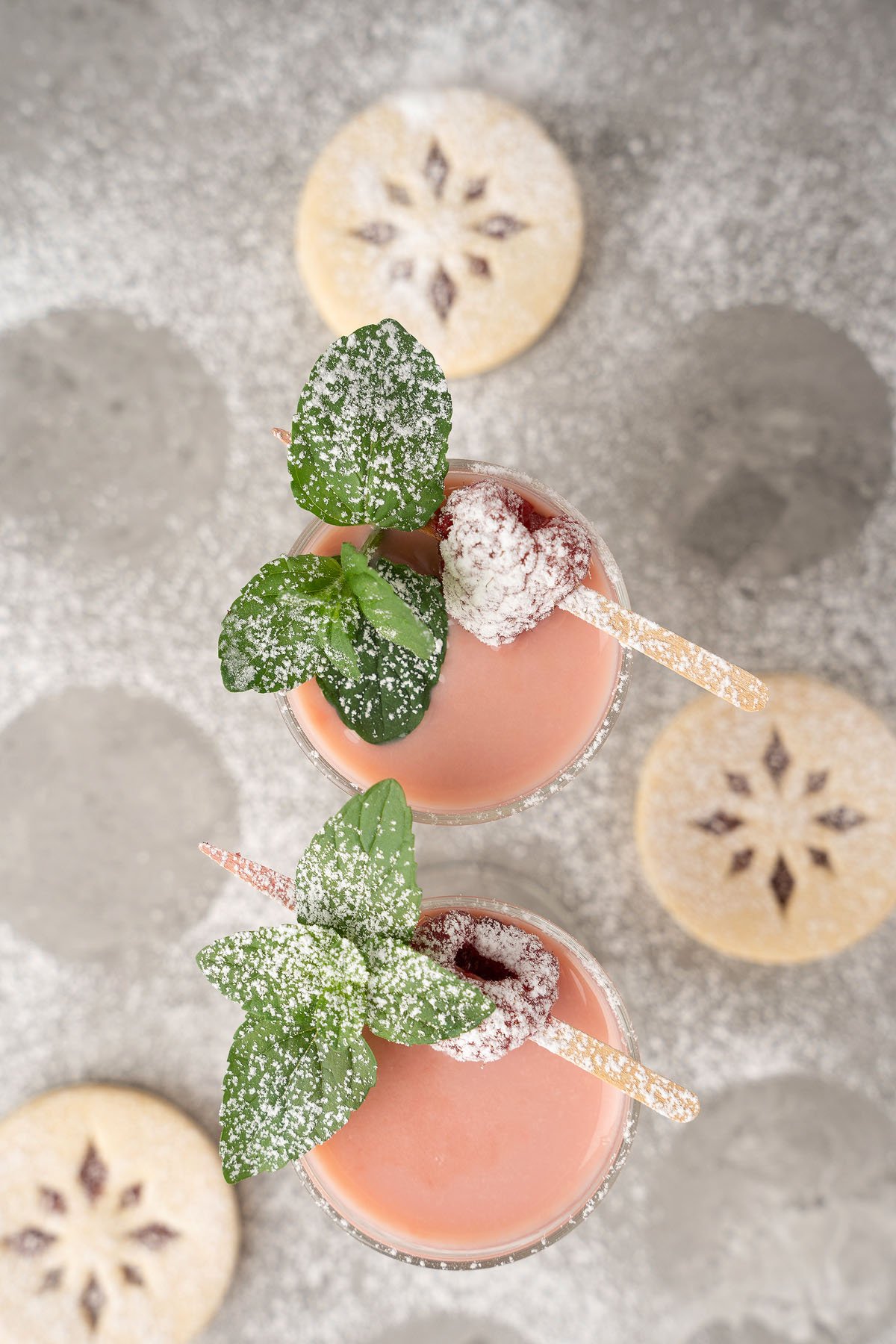 Two pink Raspberry Linzer Cookie Shots garnished with mint and berries, surrounded by round powdered sugar cookies—perfect for your next holiday cocktail or festive christmas shots recipe.