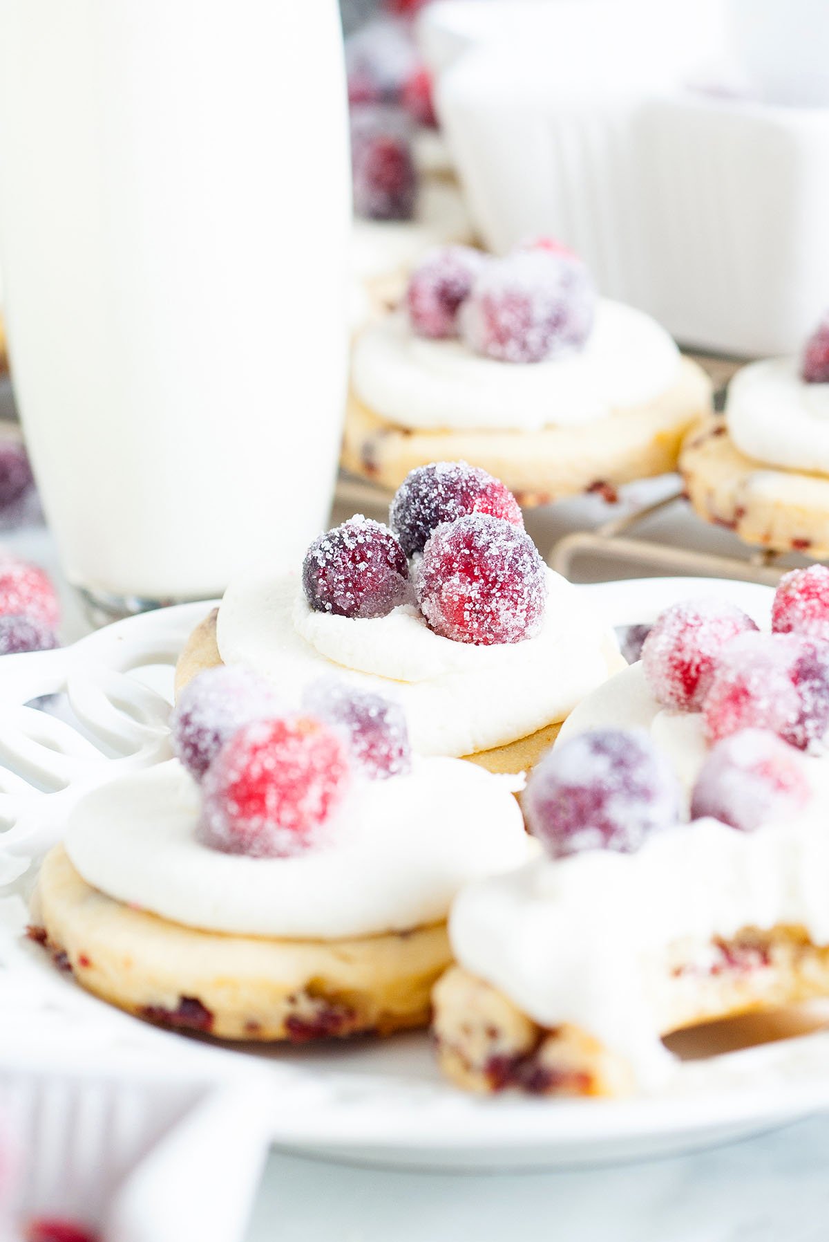 Sparkling Frosted Cranberry Cookies feature sugar cookies topped with whipped cream and sparkling cranberries, beautifully arranged on a white plate with a glass of milk.