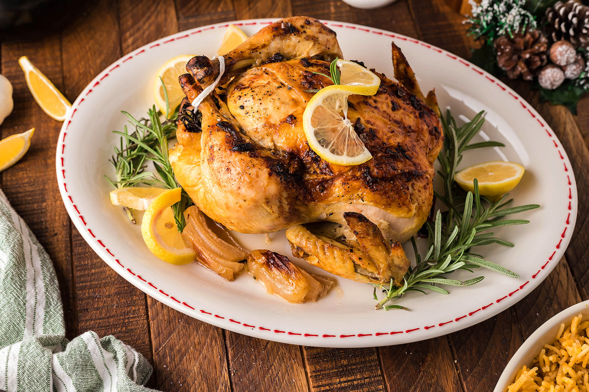 A roasted whole chicken garnished with lemon slices and fresh rosemary is placed on an oval white plate. The table setting includes a green and white striped cloth napkin beside the plate, and there are additional lemon slices arranged near the chicken.