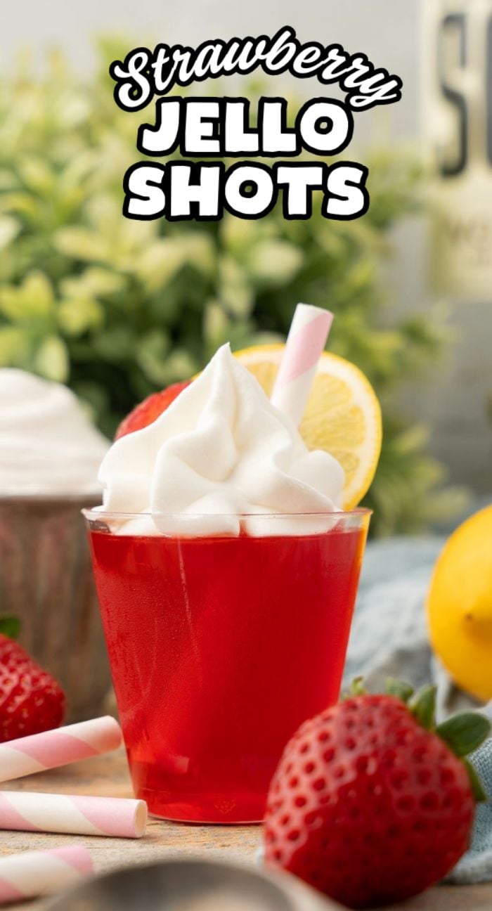 Strawberry Lemonade Jello Shots topped with whipped cream, a lemon slice, and a straw, surrounded by fresh strawberries—these boozy bites are the perfect fun treat for any party.