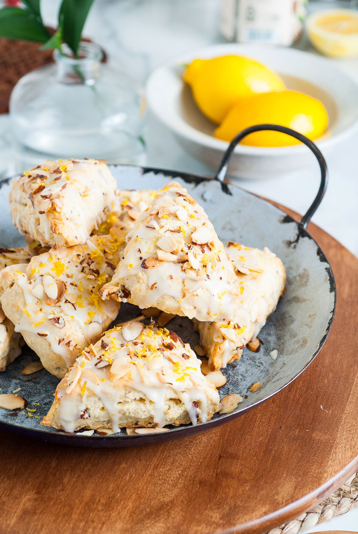 Lemon Almond Scones topped with sliced almonds and icing on a metal tray, with lemons and a vase in the background—an inviting scene for anyone seeking an easy scone recipe.