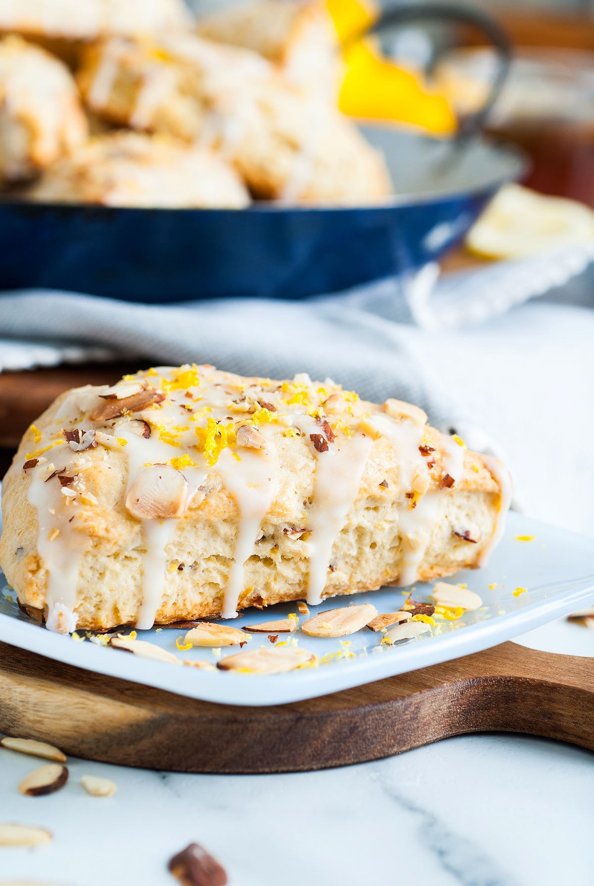 A glazed Lemon Almond Scone topped with sliced almonds and lemon zest sits on a plate, with more lemon scones in the background—a perfect treat from an easy scone recipe.