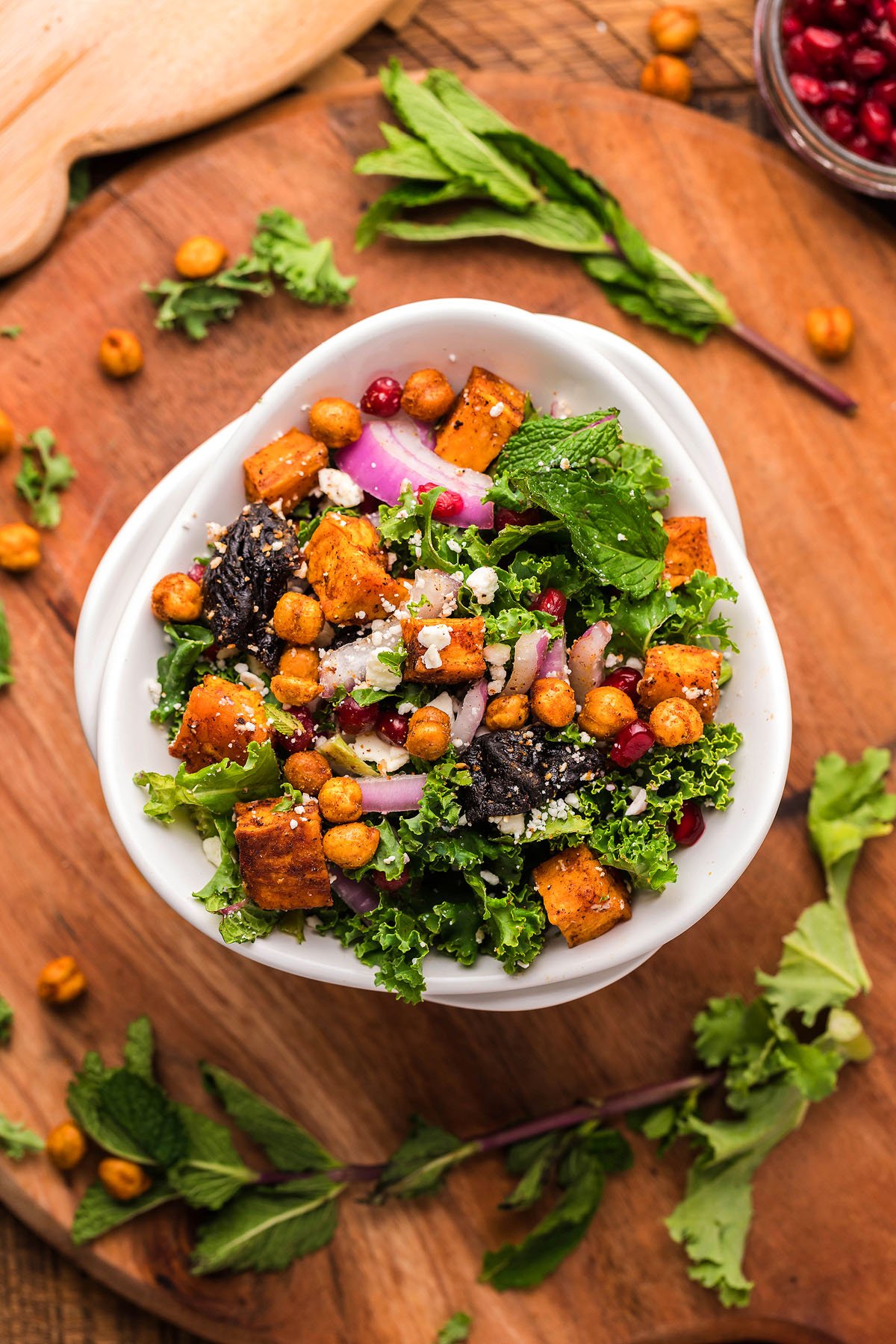 A bowl of sweet potato kale salad with roasted chickpeas, red onions, mint, and cheese sits on a wooden board.