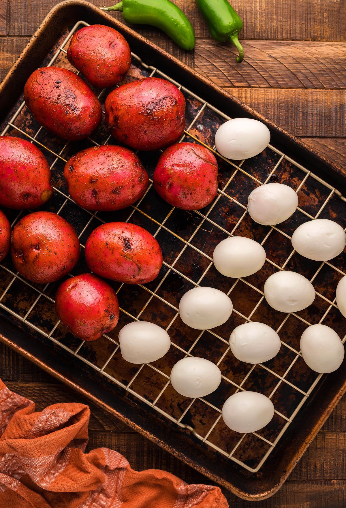 Red potatoes and white eggs on a wire rack over a baking sheet, with jalapeños and a cloth nearby—perfect for making Smoked Potato Salad with smoked potatoes and smoked hard boiled eggs on your Traeger grill or pellet smoker.