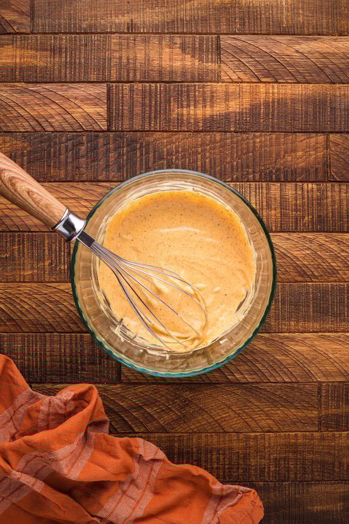 Glass bowl with creamy batter and a whisk on a wooden surface, next to an orange cloth—perfect for whipping up Smoked Potato Salad to serve alongside smoked potatoes and smoked hard boiled eggs from your Traeger grill.