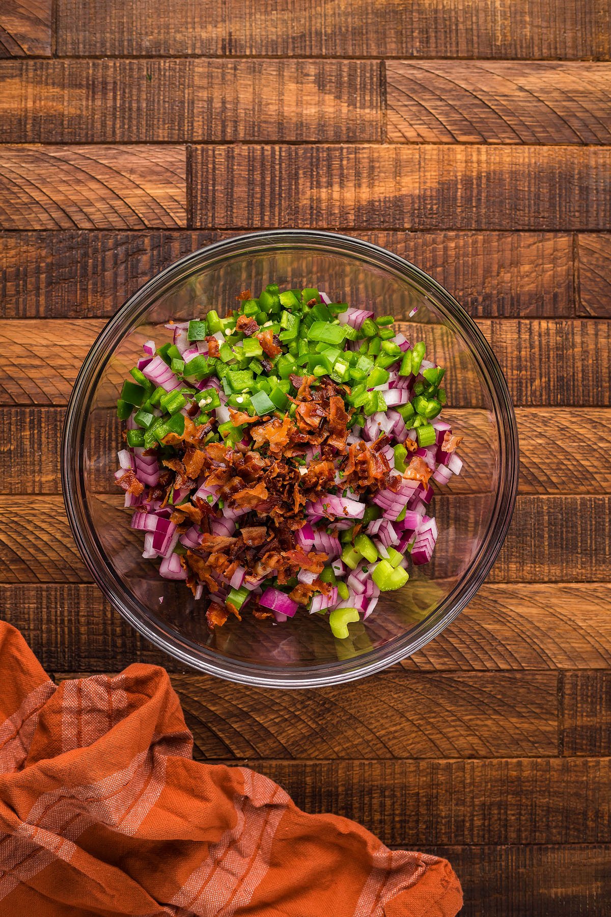 Glass bowl with chopped red onion, green pepper, celery, and bacon bits on a wooden surface—perfect for mixing up a hearty bacon potato salad to enjoy with your favorite Traeger grill or pellet smoker recipes.