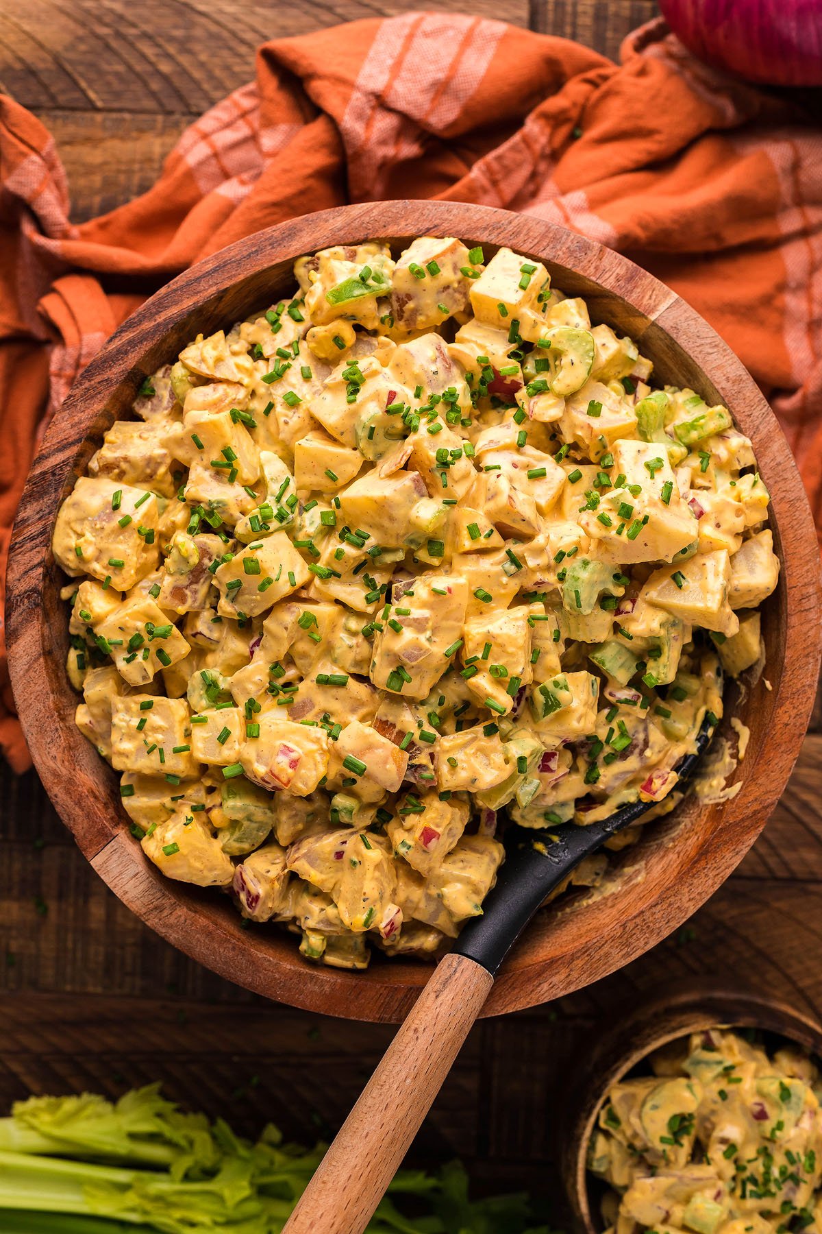 A wooden bowl filled with creamy Smoked Potato Salad, made with smoked potatoes and smoked hard boiled eggs, topped with chopped chives and served with a spoon inside. Perfect for making on your Traeger grill or pellet smoker.
