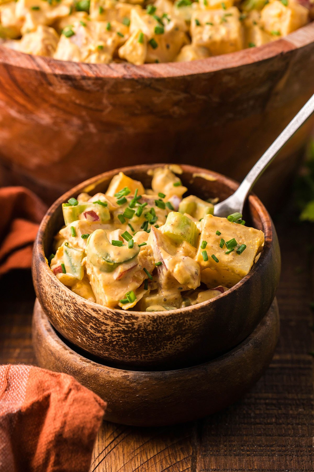 A small wooden bowl of creamy bacon potato salad garnished with chives, featuring smoked potatoes and smoked hard boiled eggs, with a larger bowl in the background.