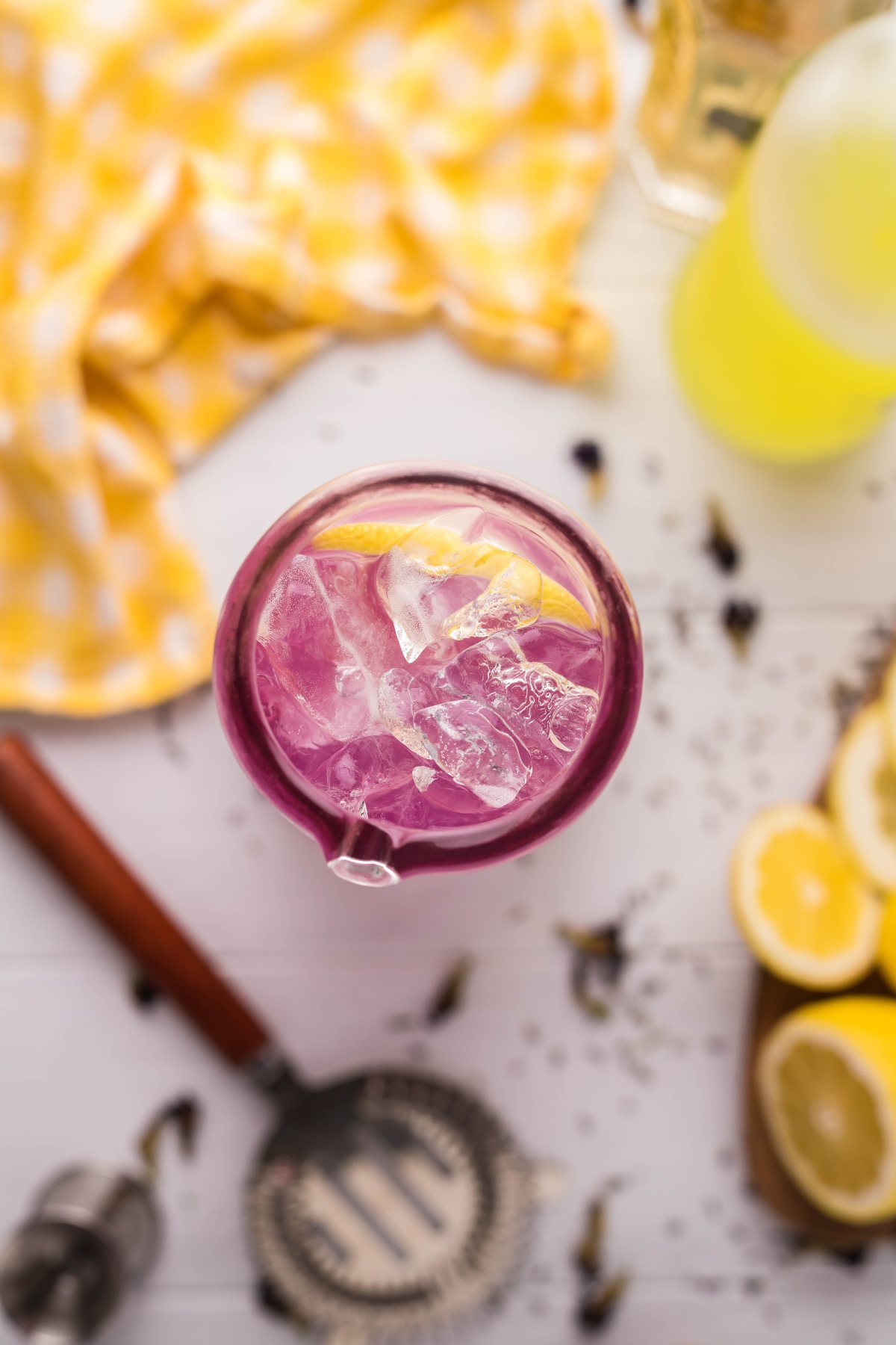 A Lemon Lavender Vodka Cocktail with ice and a lemon slice, surrounded by fresh lemons and a yellow cloth on a white table.