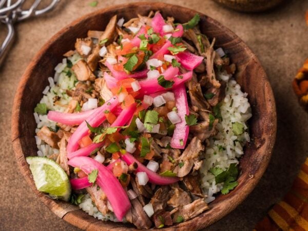 A wooden bowl of rice topped with Mexican Pulled Pork, pickled onions, fresh herbs, and a lime wedge.