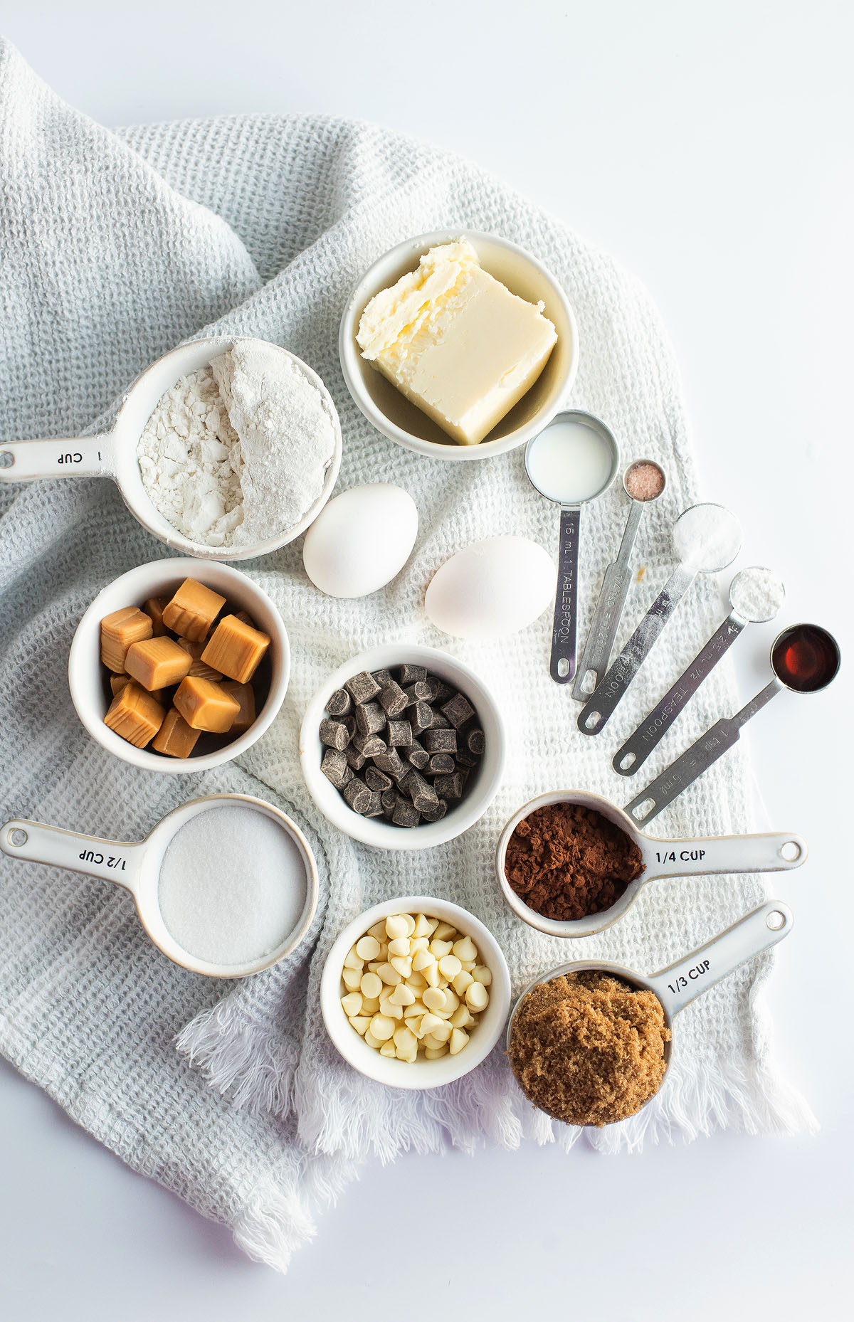 Baking ingredients in bowls and measuring cups on a knit cloth, including flour, eggs, butter, chocolate, and caramel squares for making caramel stuffed chocolate cookies.