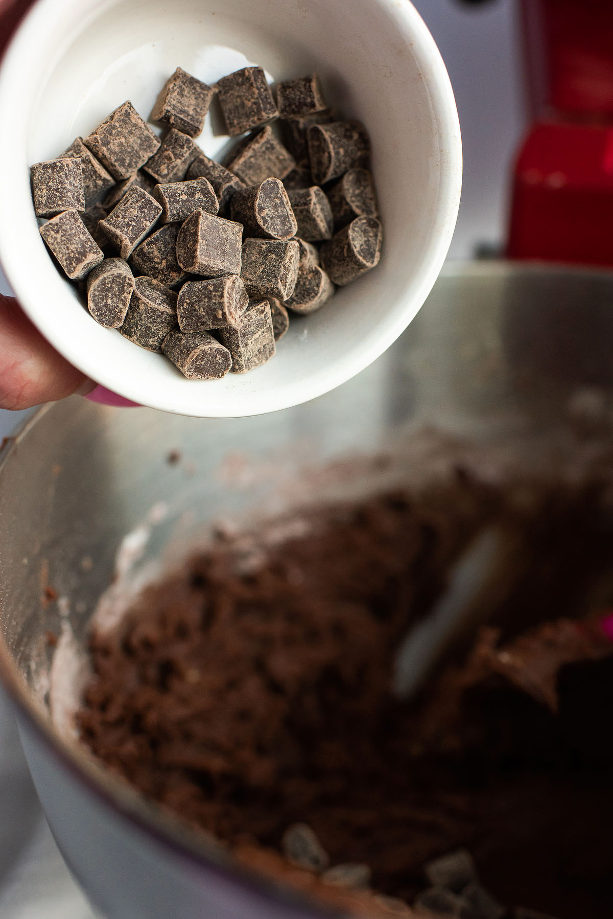 A hand holds a bowl of caramel squares above a bowl of chocolate batter in a mixer, ready to create delicious Caramel Stuffed Chocolate Cookies.