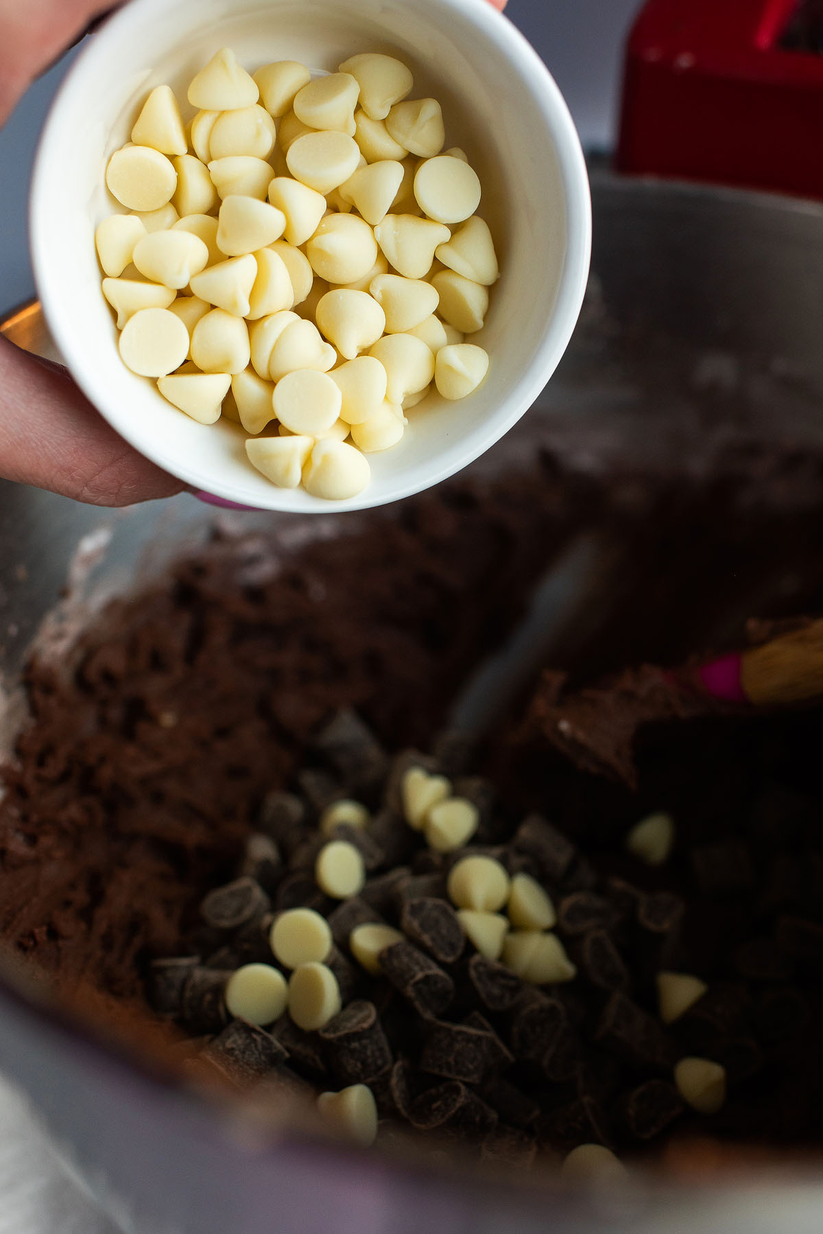 Hand holding a bowl of white chocolate chips over chocolate cookie dough dotted with dark and white chips—perfect for whipping up Caramel Stuffed Chocolate Cookies.