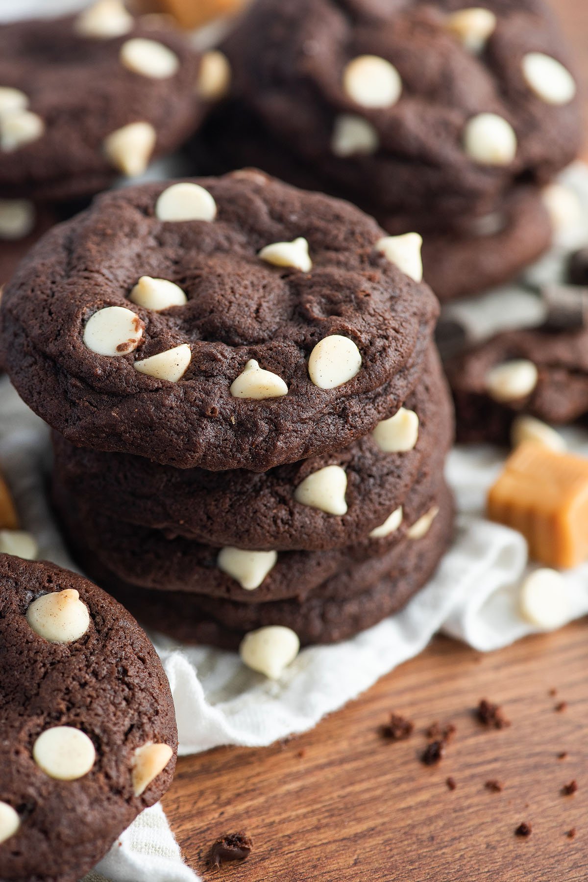 A stack of caramel stuffed chocolate cookies with white chocolate chips sits on a white cloth and wooden surface, tempting with gooey caramel centers.