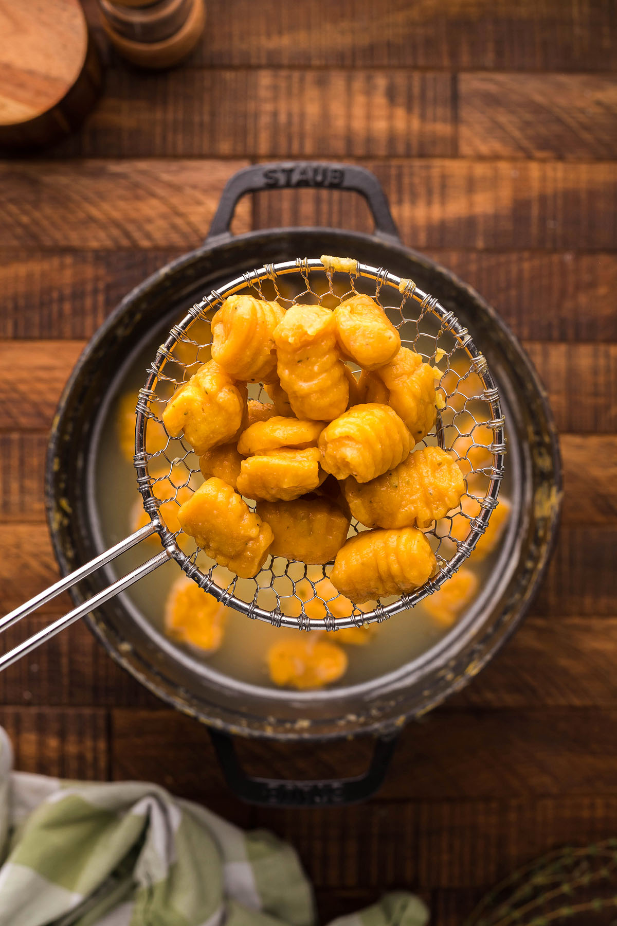 A slotted spoon holds cooked gnocchi above a pot of hot water on a wooden surface, ready for your favorite homemade potato gnocchi recipe or to be paired with a flavorful roasted garlic sauce.