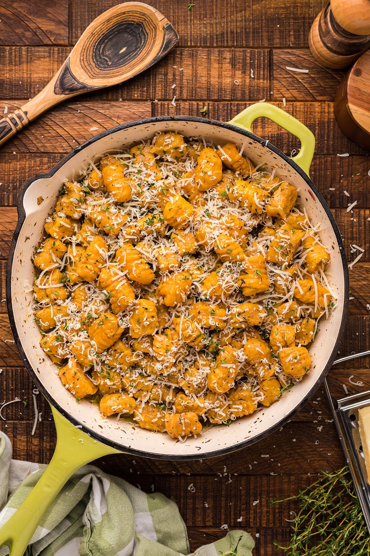 A skillet of homemade potato gnocchi topped with grated cheese and herbs sits on a wooden table, with a wooden spoon nearby.