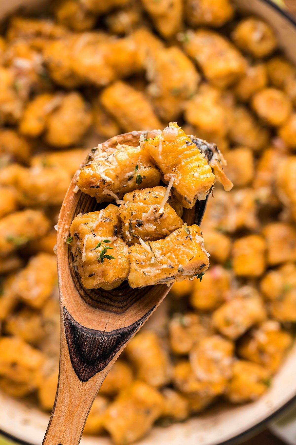 A wooden spoon holds sweet potato gnocchi topped with herbs and grated cheese above a pan full of more gnocchi, all made from a homemade potato gnocchi recipe.