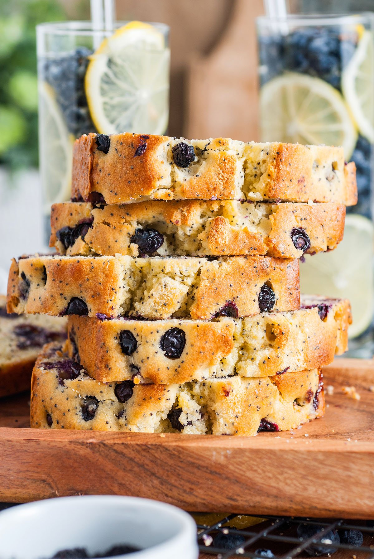 A stack of Lemon Blueberry Poppy Seed Loaf slices sits on a wooden board, with fresh lemon water in the background.