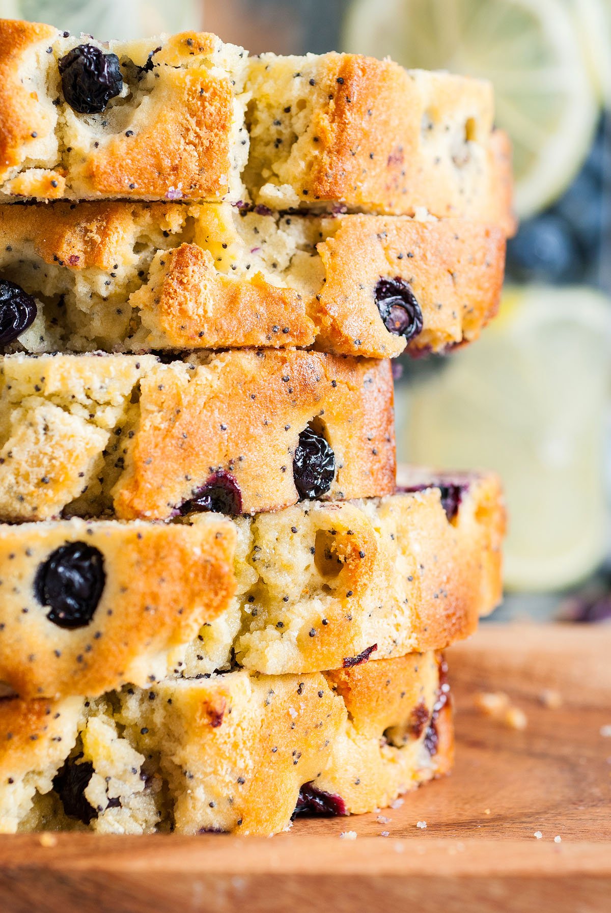 Stacked slices of Lemon Blueberry Poppy Seed Loaf on a wooden surface, with bright lemon slices in the background.