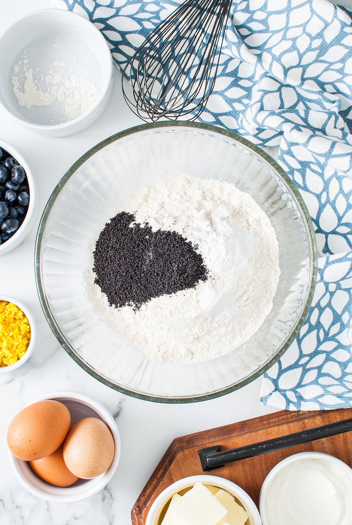 Baking ingredients in bowls, including flour, poppy seeds, eggs, butter, blueberries, and lemon zest on a counter&mdash;perfect for creating a delicious Lemon Blueberry Poppy Seed Loaf.