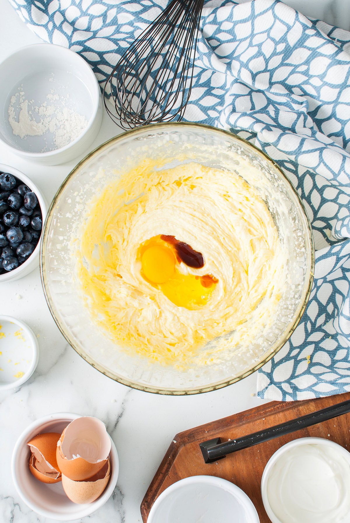 A glass bowl with butter, sugar, eggs, and vanilla being mixed to create a Lemon Blueberry Poppy Seed Loaf, surrounded by baking ingredients and utensils.