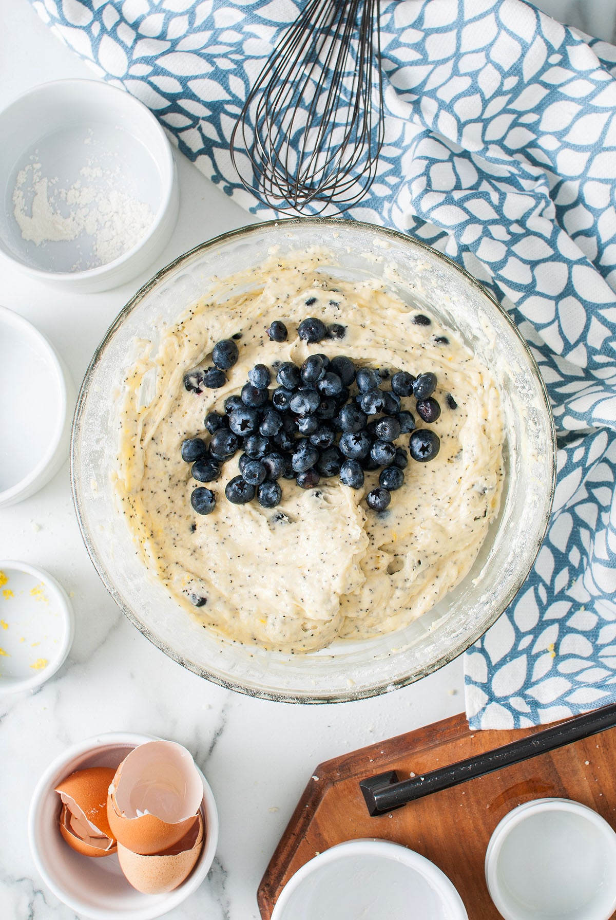 A bowl of blueberry muffin batter with fresh blueberries on top, surrounded by baking tools and eggshells&mdash;perfect for creating a Lemon Blueberry Poppy Seed Loaf at home.