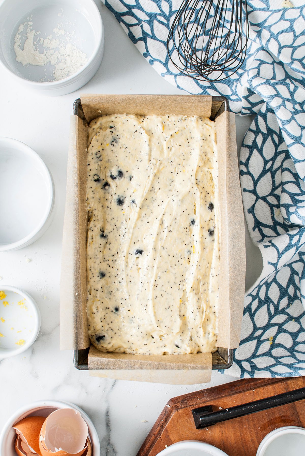 A loaf pan filled with unbaked Lemon Blueberry Poppy Seed Loaf batter, speckled with poppy seeds and blueberries, surrounded by baking tools and ingredients.