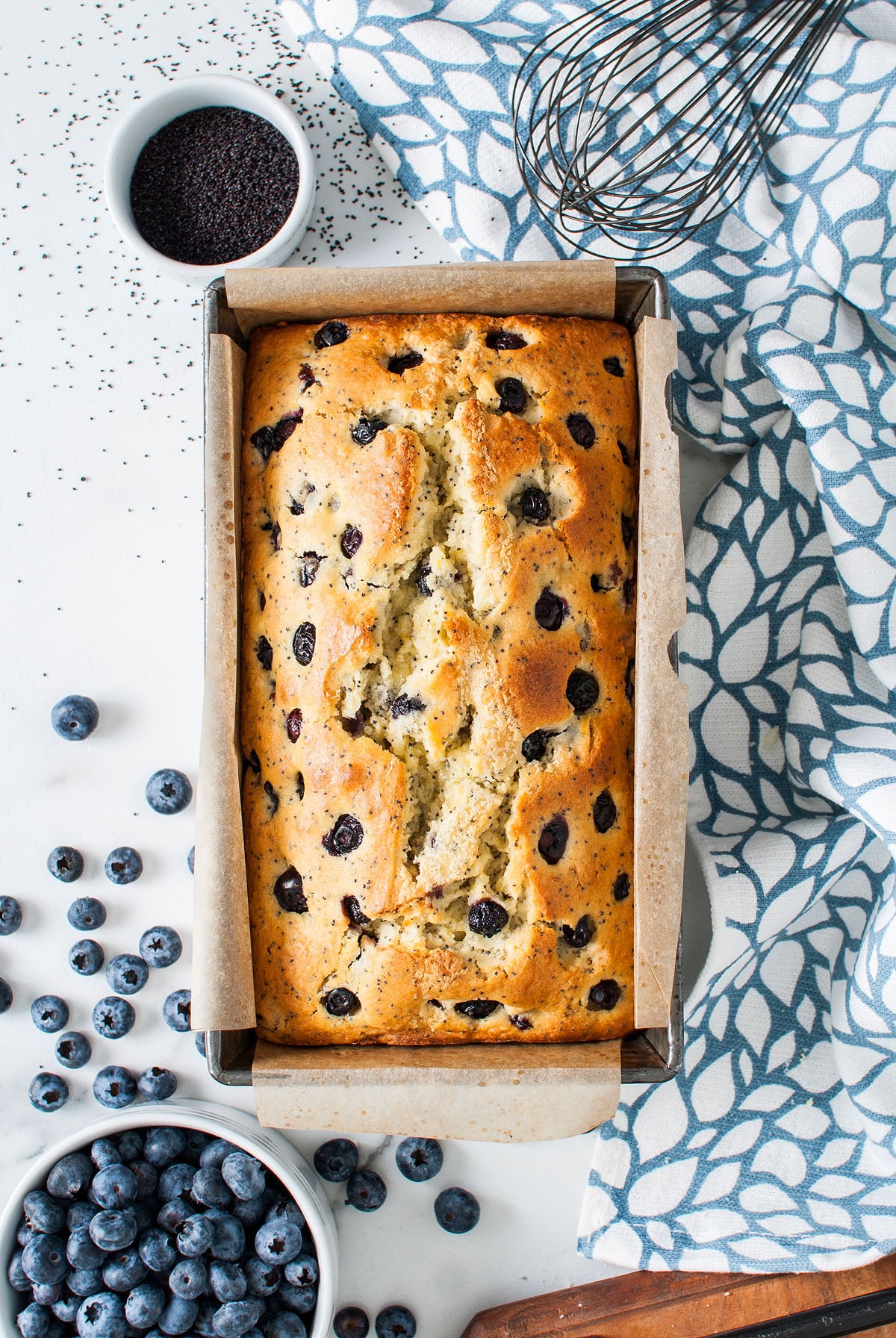 A Lemon Blueberry Poppy Seed Loaf sits in a pan, surrounded by fresh blueberries and poppy seeds on a white surface.