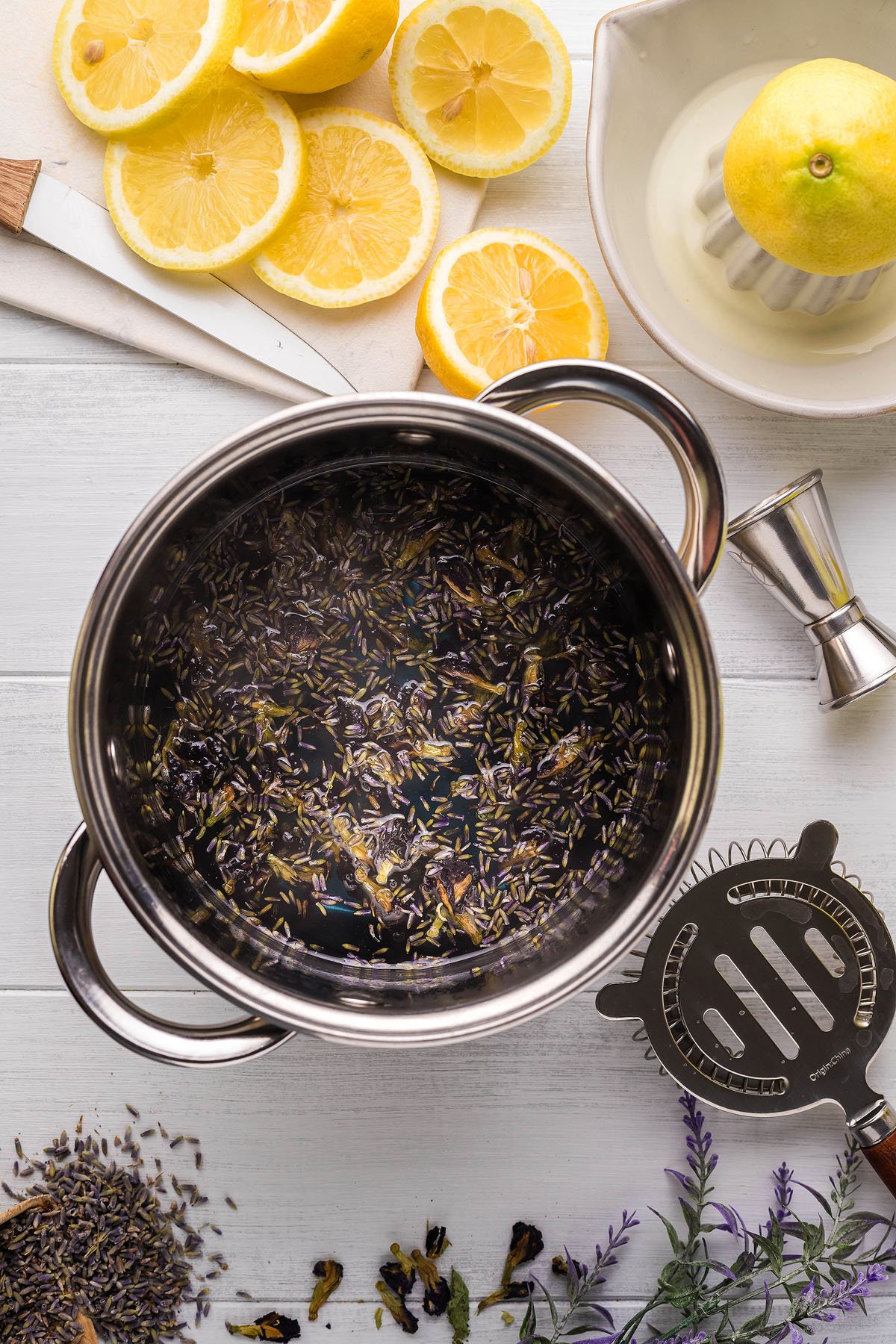 A pot with lavender and herbs next to sliced lemons, a lemon squeezer with half a lemon, and kitchen utensils on a white surface evoke the refreshing essence of a spring cocktail. Perfect for crafting a tangy lemon lavender vodka cocktail or an elegant lavender martini.