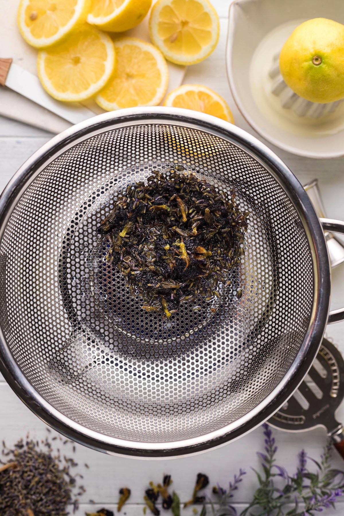 A metal strainer with loose leaf tea rests above a bowl, surrounded by lemon slices, a lemon juicer, and scattered herbs on a white surface—perfect for crafting a refreshing lemon lavender vodka cocktail.