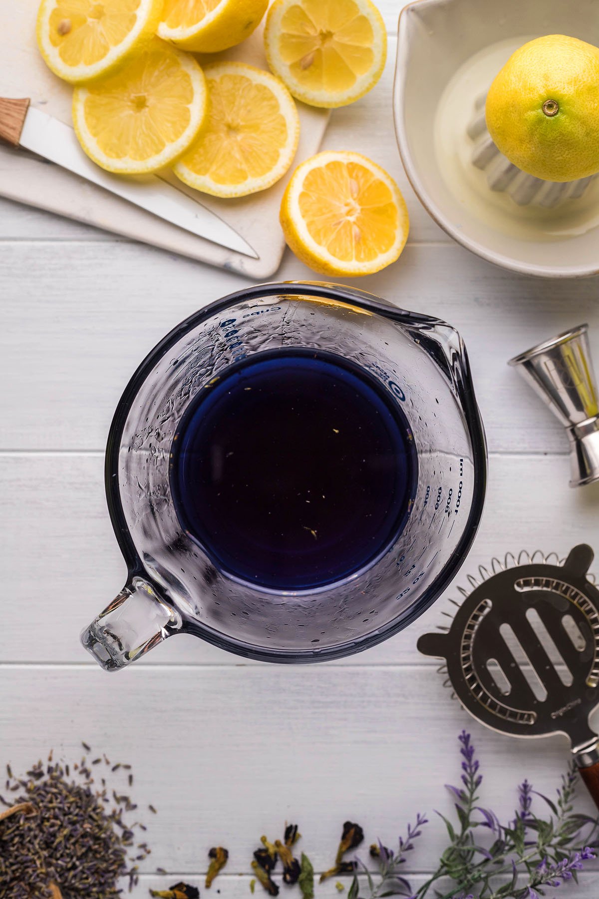 A glass pitcher with dark purple liquid sits on a white surface, embodying the essence of a Lemon Lavender Vodka Cocktail. Sliced lemons, a juicer with a lemon, a cocktail strainer, measuring jigger, and dried lavender surround it—a perfect spring cocktail setting.