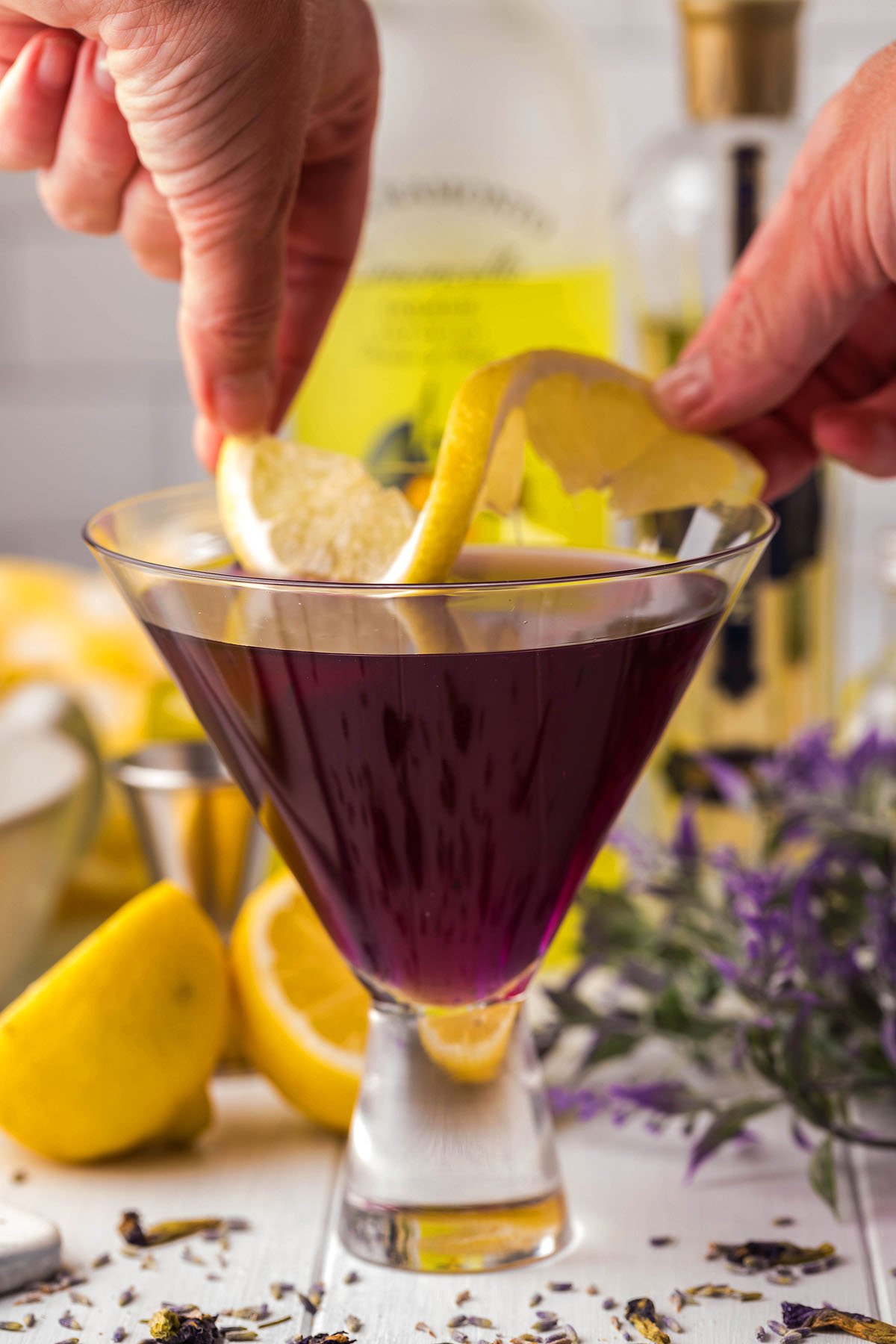 Hands delicately place a lemon slice on the rim of a purple lavender martini, set elegantly in a martini glass. In the background, fresh lemon and lavender subtly hint at the refreshing essence of this spring cocktail.