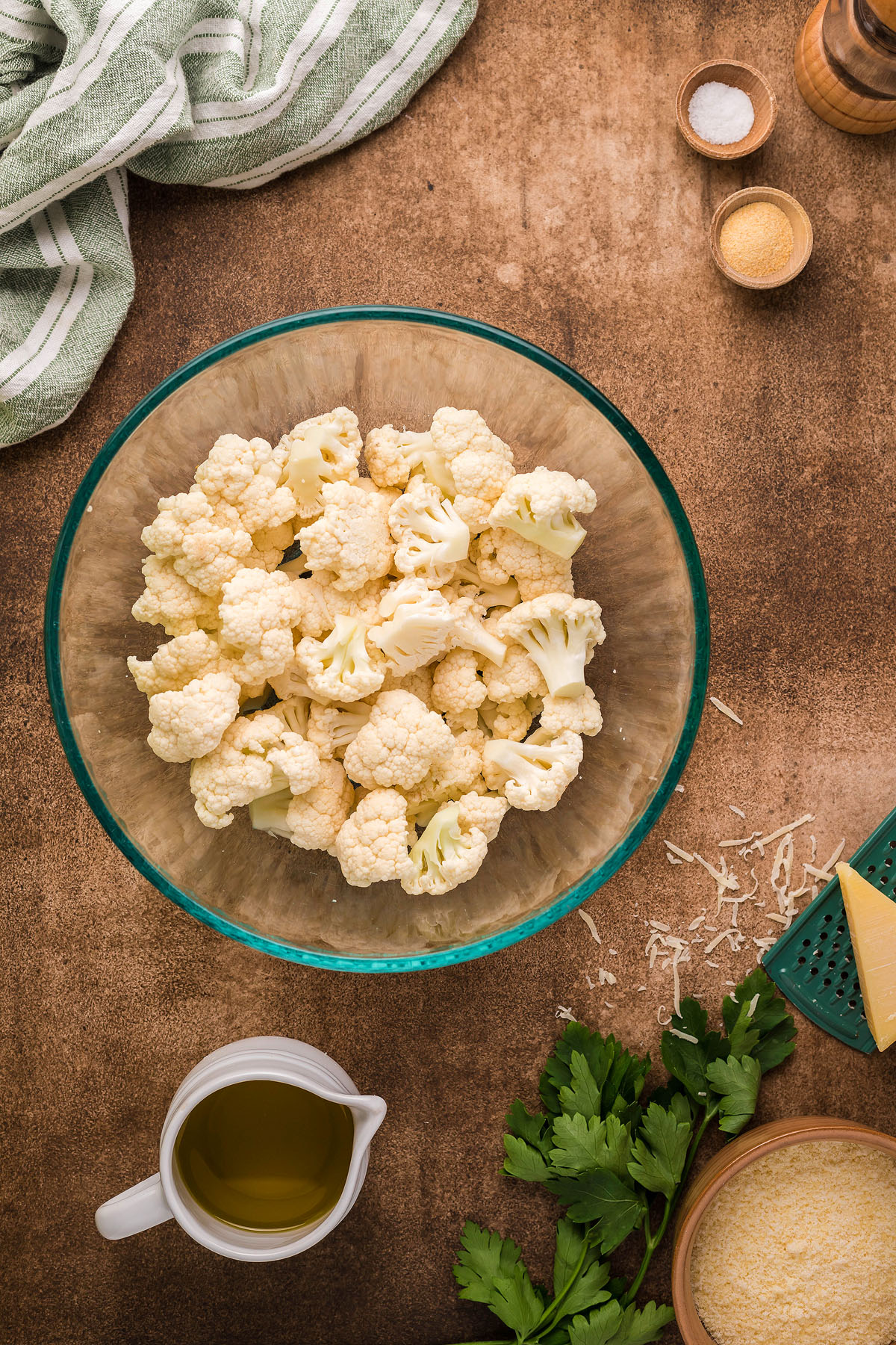 Chopped cauliflower florets in a glass bowl surrounded by oil, parsley, spices, cheese, and a striped towel—perfect for making Roasted Cauliflower Carbonara or an easy weeknight dinner with creamy cauliflower sauce.