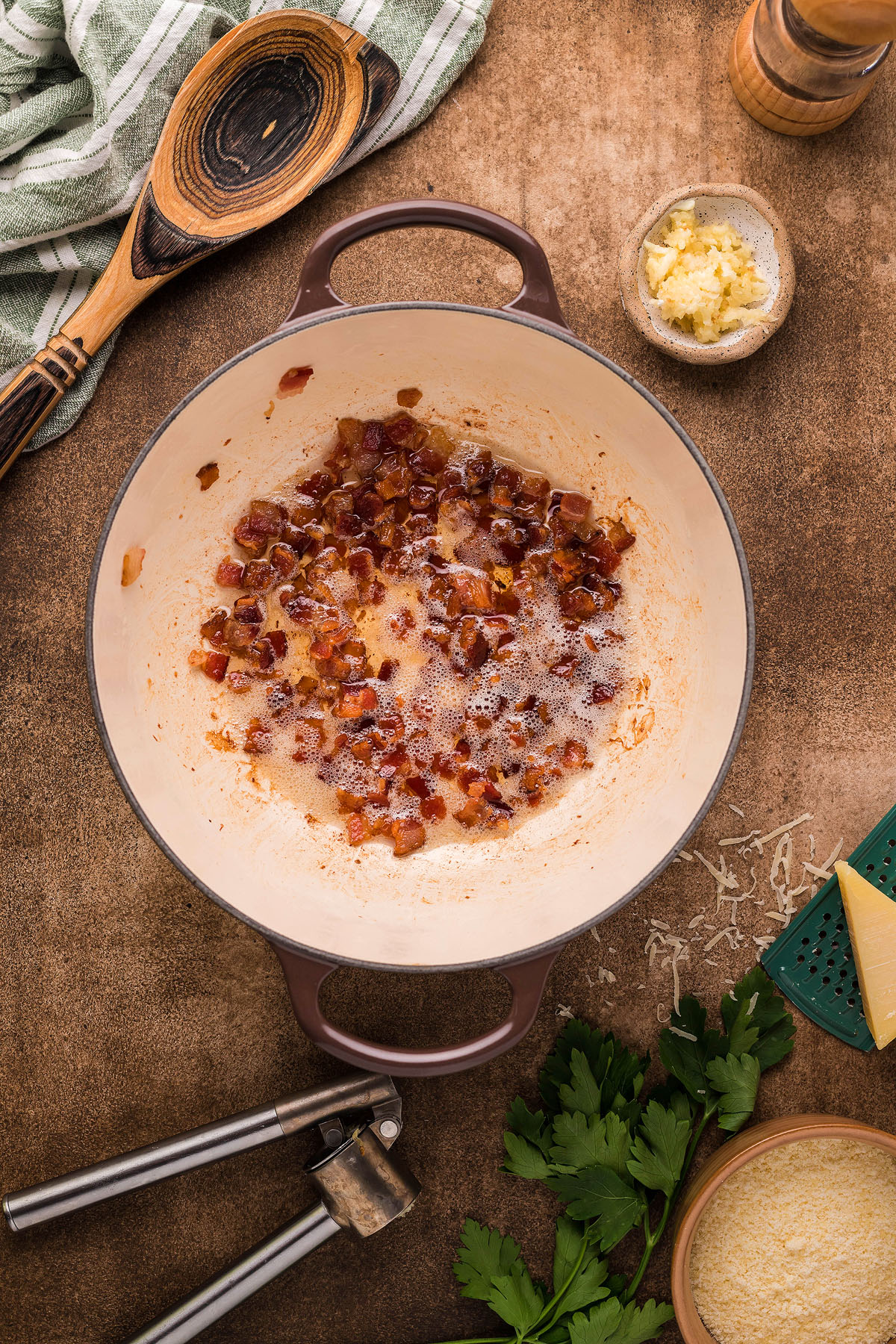 Diced bacon sizzling in a Dutch oven, surrounded by garlic, cheese, parsley, and cooking utensils—perfect for crafting Roasted Cauliflower Carbonara or an easy weeknight dinner with rich cauliflower sauce.